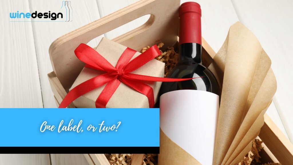 A wine bottle with a blank label packed alongside a gift box tied with a red ribbon inside a wooden crate.
